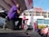 A photographer wearing a purple shirt kneels on stage taking photos at an outdoor event. A person dressed in pink, possibly a performer or speaker, stands on stage holding a pink ribbon, while a crowd of people in pink attire watches. Balloons and stage equipment are visible, with a large building and clear blue sky in the background.