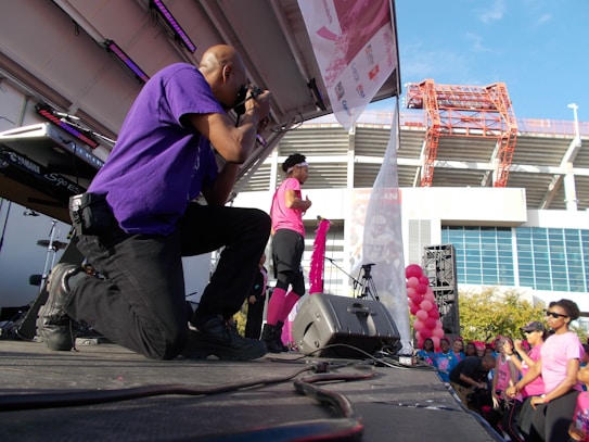 A photographer wearing a purple shirt kneels on stage taking photos at an outdoor event. A person dressed in pink, possibly a performer or speaker, stands on stage holding a pink ribbon, while a crowd of people in pink attire watches. Balloons and stage equipment are visible, with a large building and clear blue sky in the background.