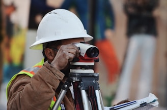 A person wearing a white hard hat and high-visibility vest operates a surveying instrument, focusing intently on the task at hand. The background is blurred with hints of other construction activities or team members.
