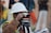 A person wearing a white hard hat and high-visibility vest operates a surveying instrument, focusing intently on the task at hand. The background is blurred with hints of other construction activities or team members.