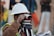 A person wearing a white hard hat and high-visibility vest operates a surveying instrument, focusing intently on the task at hand. The background is blurred with hints of other construction activities or team members.