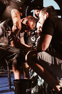 A group of people intensely focus on an activity involving one person in the center, possibly providing support or guidance. The setting appears to be indoors with a cage or netted area in the background, hinting at a sports or competitive environment. The lighting creates dramatic shadows, emphasizing the concentration and seriousness of the moment.