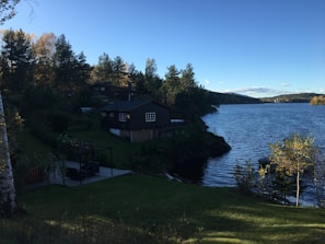 A serene lakeside landscape with a wooden cabin situated near the water's edge, surrounded by lush green trees. The calm water reflects the clear blue sky, while the backdrop includes a mix of dense forest and distant hills. A well-maintained grassy area leads towards the cabin, creating a peaceful and natural setting.