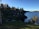 A serene lakeside landscape with a wooden cabin situated near the water's edge, surrounded by lush green trees. The calm water reflects the clear blue sky, while the backdrop includes a mix of dense forest and distant hills. A well-maintained grassy area leads towards the cabin, creating a peaceful and natural setting.