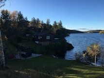A serene lakeside landscape with a wooden cabin situated near the water's edge, surrounded by lush green trees. The calm water reflects the clear blue sky, while the backdrop includes a mix of dense forest and distant hills. A well-maintained grassy area leads towards the cabin, creating a peaceful and natural setting.