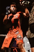 A male mixed martial arts fighter is in action, wearing bright red shorts and a matching shirt with various logos. His stance suggests he is either defending or preparing to attack. The fight appears to be taking place inside a caged arena, with a referee visible in the background.