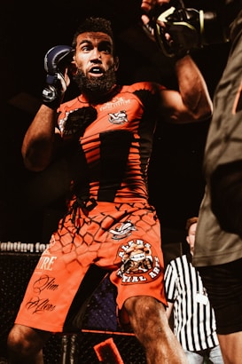 A male mixed martial arts fighter is in action, wearing bright red shorts and a matching shirt with various logos. His stance suggests he is either defending or preparing to attack. The fight appears to be taking place inside a caged arena, with a referee visible in the background.