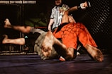 Two individuals are engaged in a mixed martial arts match inside a cage. One person wearing red shorts is pinning the other dressed in grey shorts to the mat. A referee in a black and white striped shirt observes the match closely.