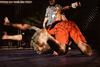 Two individuals are engaged in a mixed martial arts match inside a cage. One person wearing red shorts is pinning the other dressed in grey shorts to the mat. A referee in a black and white striped shirt observes the match closely.