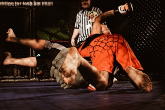 Two individuals are engaged in a mixed martial arts match inside a cage. One person wearing red shorts is pinning the other dressed in grey shorts to the mat. A referee in a black and white striped shirt observes the match closely.