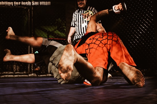 Two individuals are engaged in a mixed martial arts match inside a cage. One person wearing red shorts is pinning the other dressed in grey shorts to the mat. A referee in a black and white striped shirt observes the match closely.
