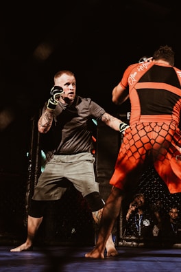 Two fighters are engaged in a mixed martial arts match inside a caged arena. One fighter is wearing a black and gray outfit with protective gloves, while the other is dressed in red and black. The audience is partially visible in the background, watching intently.