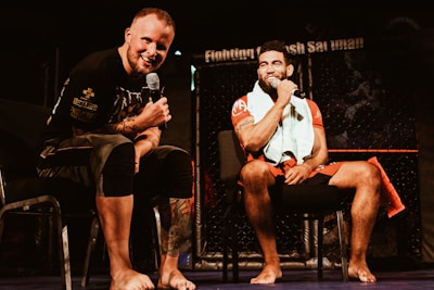 Two individuals are seated on a stage, each holding a microphone and smiling. They are in a sports-related setting with one wearing a casual black outfit and the other in sportswear, with a towel around his neck. The background includes elements of a fighting or martial arts cage.