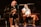 Two individuals are seated on a stage, each holding a microphone and smiling. They are in a sports-related setting with one wearing a casual black outfit and the other in sportswear, with a towel around his neck. The background includes elements of a fighting or martial arts cage.