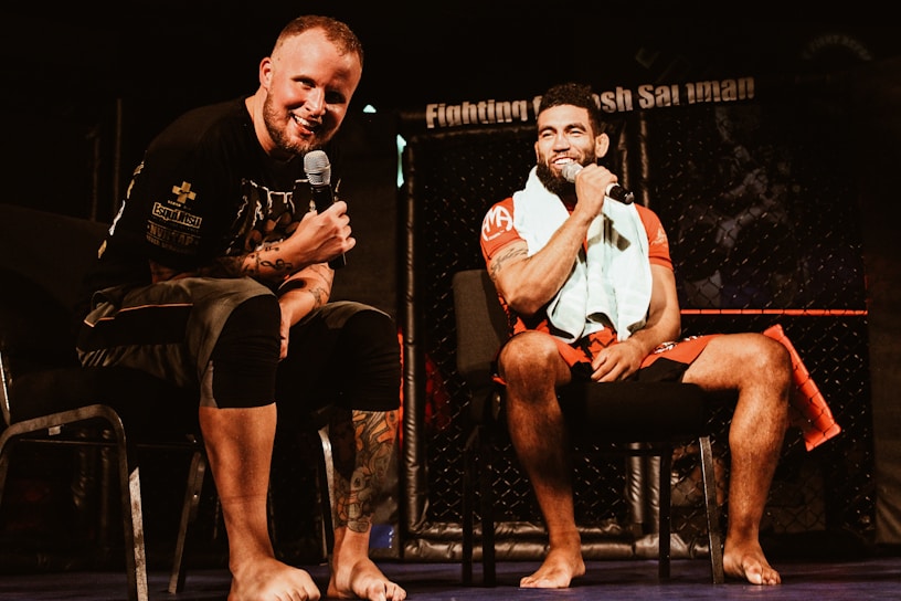 Two individuals are seated on a stage, each holding a microphone and smiling. They are in a sports-related setting with one wearing a casual black outfit and the other in sportswear, with a towel around his neck. The background includes elements of a fighting or martial arts cage.