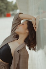 A person leans back against a wall with an arm resting over their head, eyes closed, conveying a sense of relaxation or introspection. The backdrop features a blurred outdoor scene with muted colors.