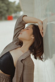 A person leans back against a wall with an arm resting over their head, eyes closed, conveying a sense of relaxation or introspection. The backdrop features a blurred outdoor scene with muted colors.