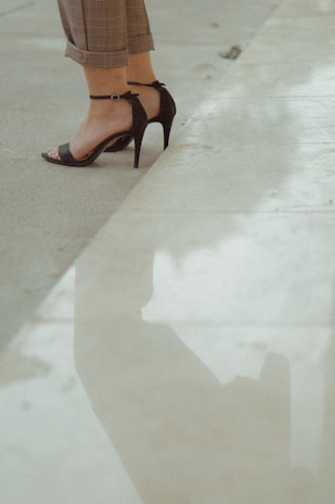 High-heeled sandals on bare feet are partially visible with cropped trousers. The person is standing next to a reflective surface, likely water or polished stone, creating a faint reflection of the lower legs and shoes.