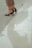 High-heeled sandals on bare feet are partially visible with cropped trousers. The person is standing next to a reflective surface, likely water or polished stone, creating a faint reflection of the lower legs and shoes.