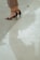 High-heeled sandals on bare feet are partially visible with cropped trousers. The person is standing next to a reflective surface, likely water or polished stone, creating a faint reflection of the lower legs and shoes.