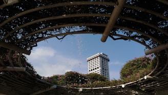 A modern high-rise building is framed by a circular, open pergola structure covered in lush greenery and flowers. Water droplets from a fountain arc gracefully through the air against a clear blue sky.
