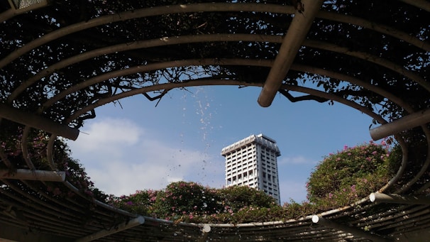 A modern high-rise building is framed by a circular, open pergola structure covered in lush greenery and flowers. Water droplets from a fountain arc gracefully through the air against a clear blue sky.
