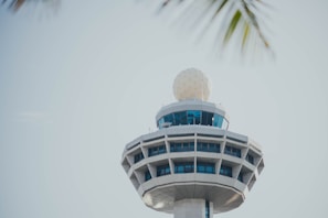 A modern air traffic control tower with a spherical radar dome on top, featuring large windows and an angular design. The sky is clear and slightly overcast.