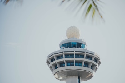 A modern air traffic control tower with a spherical radar dome on top, featuring large windows and an angular design. The sky is clear and slightly overcast.
