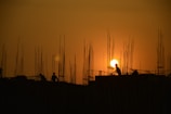 Silhouettes of construction workers are visible against a dramatic orange sunset sky, with numerous metal rods protruding from the structure they are working on. The sun appears prominently between the rods, creating a visually striking scene.