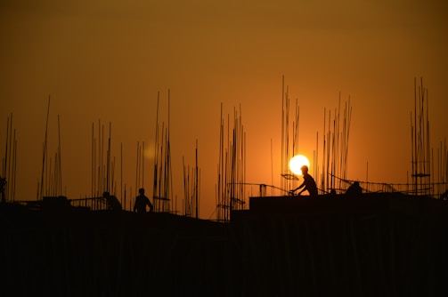 Silhouettes of construction workers are visible against a dramatic orange sunset sky, with numerous metal rods protruding from the structure they are working on. The sun appears prominently between the rods, creating a visually striking scene.