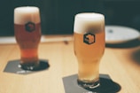 Two glasses of beer with thick foam heads sitting on dark coasters on a wooden table. The glass branding reads 'SVB Spring Valley Brewery'. The background is softly blurred, giving the setting a calm and cozy atmosphere.