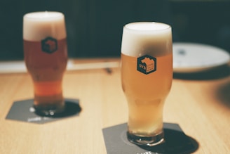 Two glasses of beer with thick foam heads sitting on dark coasters on a wooden table. The glass branding reads 'SVB Spring Valley Brewery'. The background is softly blurred, giving the setting a calm and cozy atmosphere.