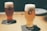 Two glasses of beer with thick foam heads sitting on dark coasters on a wooden table. The glass branding reads 'SVB Spring Valley Brewery'. The background is softly blurred, giving the setting a calm and cozy atmosphere.