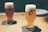 Two glasses of beer with thick foam heads sitting on dark coasters on a wooden table. The glass branding reads 'SVB Spring Valley Brewery'. The background is softly blurred, giving the setting a calm and cozy atmosphere.