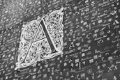 A large, ornate letter 'A' is prominently displayed in a decorative style with intricate floral and scroll patterns. Surrounding the letter are numerous smaller symbols, letters, and scripts in various fonts and styles, resembling a collage or collection of calligraphy samples. The image is monochromatic, with shades of gray and white.
