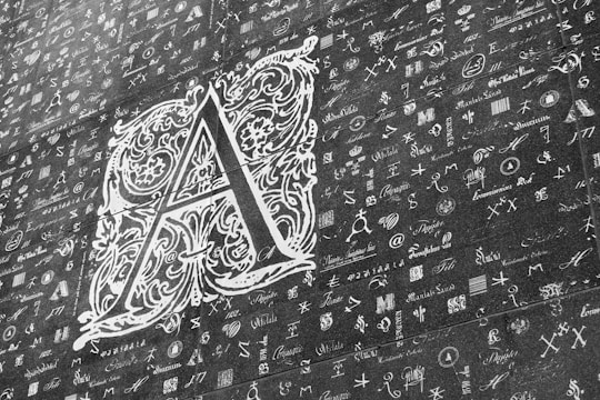A large, ornate letter 'A' is prominently displayed in a decorative style with intricate floral and scroll patterns. Surrounding the letter are numerous smaller symbols, letters, and scripts in various fonts and styles, resembling a collage or collection of calligraphy samples. The image is monochromatic, with shades of gray and white.
