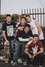 Five young men are gathered in a casual outdoor setting. Four of them are standing, and one is crouching in front. They are dressed in casual clothing, including graphic t-shirts and jeans. One person is holding a skateboard, and all of them appear to be smiling or laughing, suggesting a lighthearted atmosphere. A fence and some greenery are visible in the background.