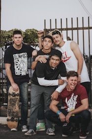 Five young men are gathered in a casual outdoor setting. Four of them are standing, and one is crouching in front. They are dressed in casual clothing, including graphic t-shirts and jeans. One person is holding a skateboard, and all of them appear to be smiling or laughing, suggesting a lighthearted atmosphere. A fence and some greenery are visible in the background.
