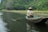 A person wearing a traditional conical hat is rowing a small boat through calm water, surrounded by lush green vegetation and rocky cliffs.