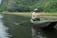 A person wearing a traditional conical hat is rowing a small boat through calm water, surrounded by lush green vegetation and rocky cliffs.