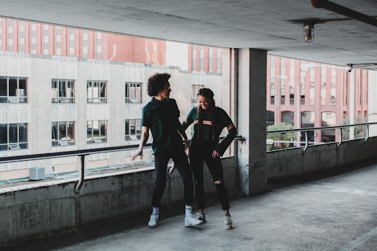 Two people stand in an urban parking garage, with a cityscape of buildings visible in the background. They both appear to be casually dressed and are smiling at each other, conveying a playful interaction.