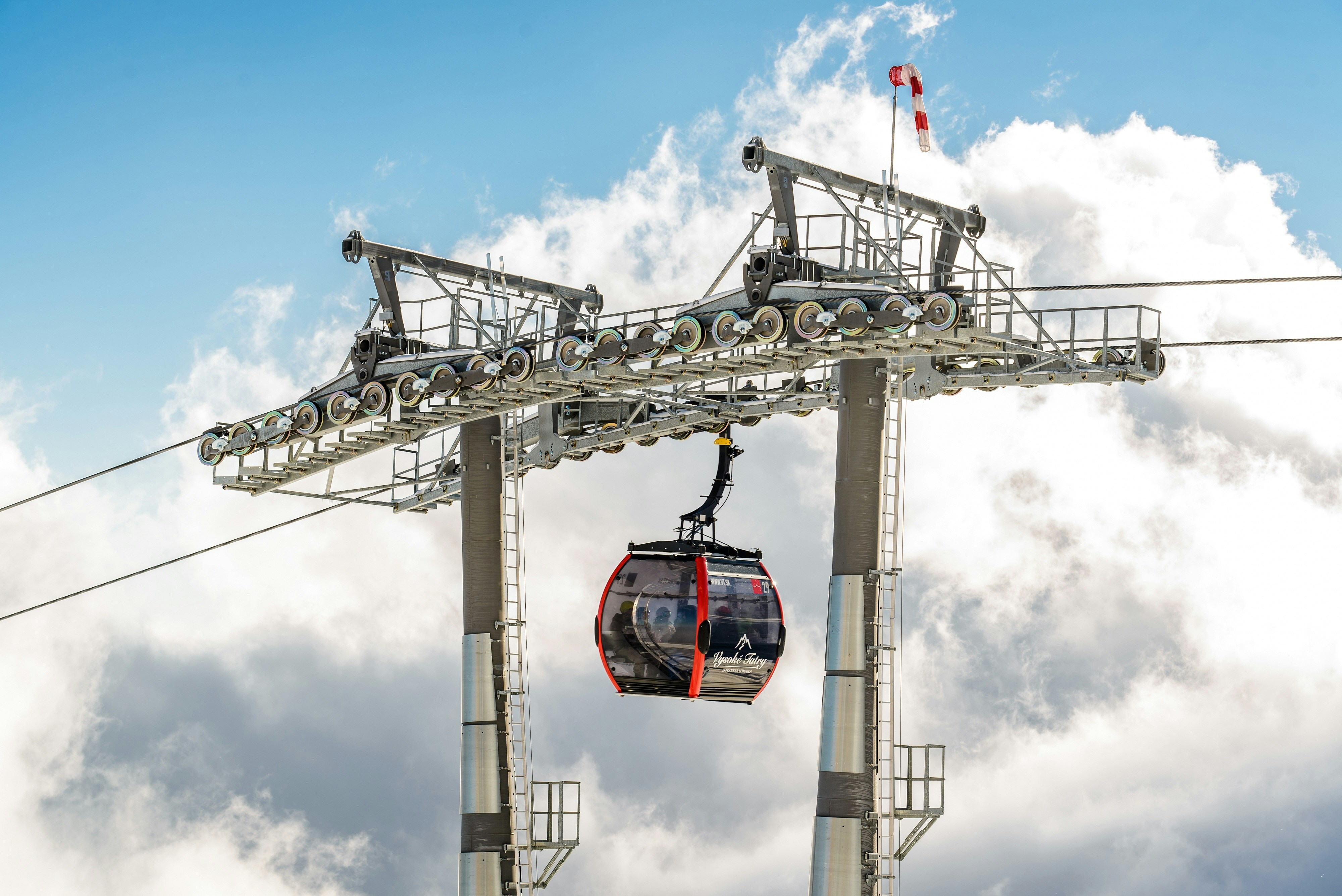 A gondola lift suspended from a cable glides near a support tower against a backdrop of bright blue sky and fluffy white clouds. The gondola is black and red with large windows, allowing a view of the interior. The structural elements of the lift are metallic, reinforcing its robust construction.