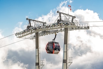 A gondola lift suspended from a cable glides near a support tower against a backdrop of bright blue sky and fluffy white clouds. The gondola is black and red with large windows, allowing a view of the interior. The structural elements of the lift are metallic, reinforcing its robust construction.