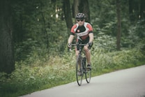 A cyclist wearing a black helmet, sunglasses, and a red and white cycling jersey is riding a black road bike on a paved path through a green forest. Vegetation surrounds the cyclist, and tall trees frame the background.