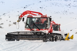 Two red snow grooming machines are parked on a snowy landscape with mountains in the background. The snowcats have large black front blades and are equipped with cabin lights. The snow around the machines is pristine and undisturbed.