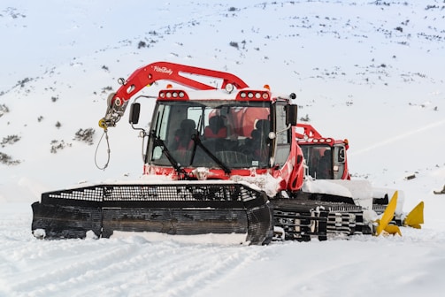 Two red snow grooming machines are parked on a snowy landscape with mountains in the background. The snowcats have large black front blades and are equipped with cabin lights. The snow around the machines is pristine and undisturbed.