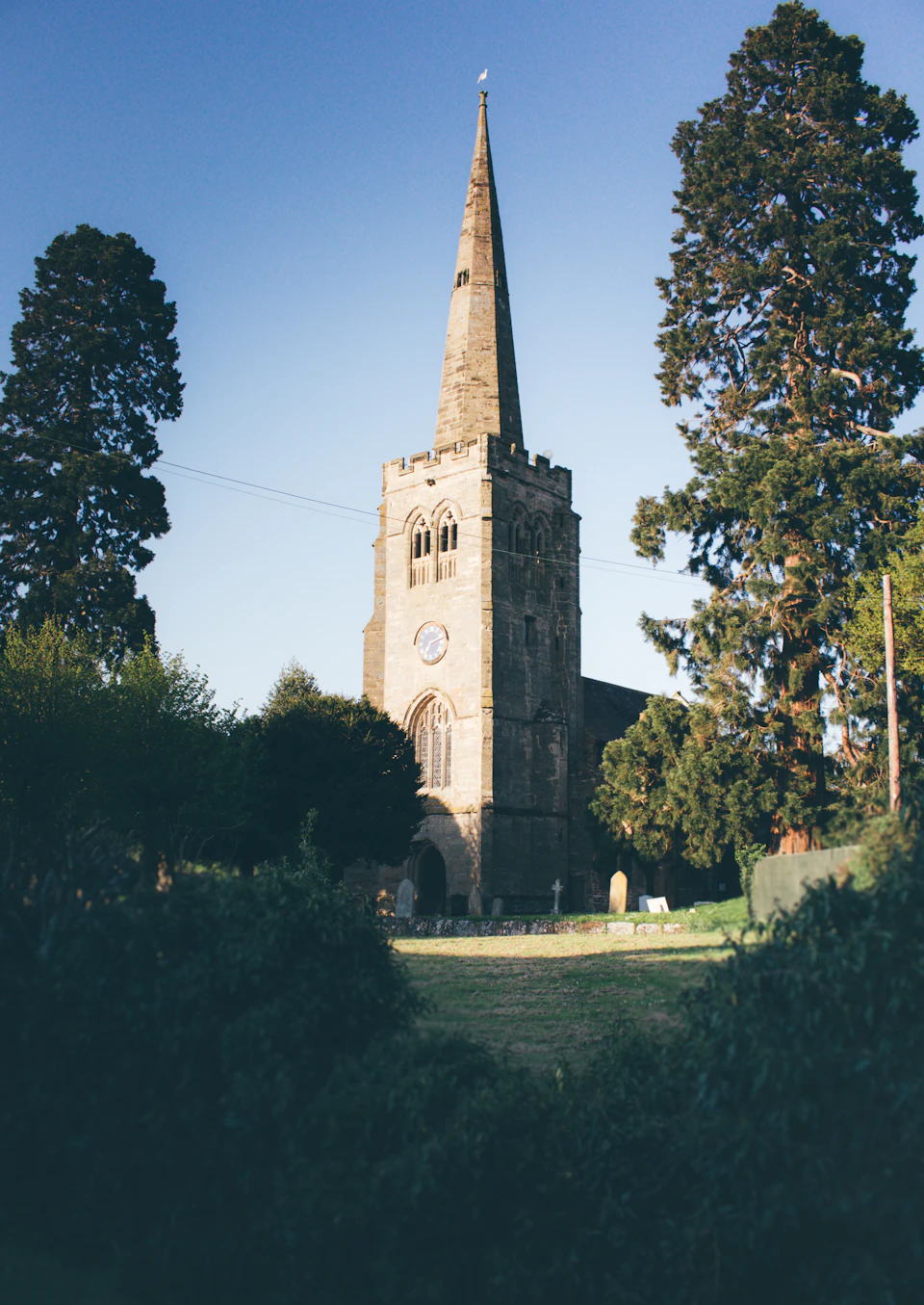 The image depicts a serene scene at a church, with its tall steeple reaching towards the clear blue sky. The church is nestled amidst lush greenery, including trees and bushes that surround it on all sides. In front of the church, there's a small cemetery, adding to the peaceful atmosphere of the location.