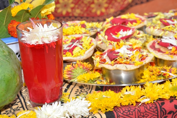 A vibrant arrangement of festive items, featuring a glass filled with a red liquid and white flowers submerged on top. Surrounding the glass are several dishes containing food adorned with red color powder and yellow marigold petals. These items are arranged on an intricately patterned fabric, conveying a sense of celebration.