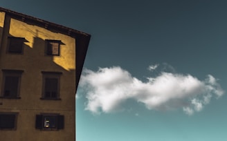 A single cloud is floating in a clear sky next to a building with yellowish walls and dark window shutters. The building occupies the left side of the image, casting subtle shadows.