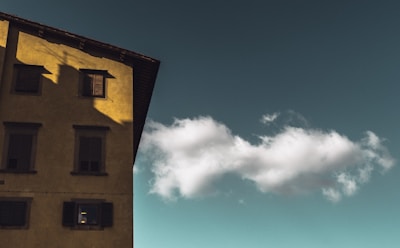 A single cloud is floating in a clear sky next to a building with yellowish walls and dark window shutters. The building occupies the left side of the image, casting subtle shadows.